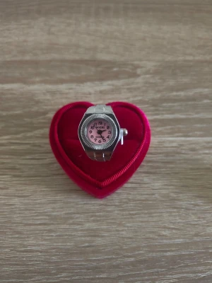 Silverfärgad ringklocka med rosa urtavla - Unik ringklocka från Quartz i silverfärg med elastiskt band och rund rosa urtavla. Siffrorna är tydliga och boetten har en stilren, modern look. Perfekt accessoar för dig som vill ha något annorlunda på fingret.