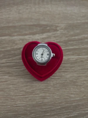 Silverfärgad ring med klockmotiv - Unik silverfärgad ring utformad som en klassisk klocka med vit urtavla och romerska siffror. Ringen har en bred design och detaljerad urtavla, vilket ger en cool och retro känsla. Perfekt accessoar för dig som gillar statement-smycken.