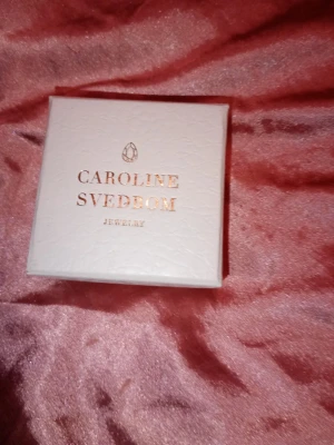 Smyckesask Caroline Svedbom - Vit fyrkantig smyckesask från Caroline Svedbom med guldig logotyp på locket. Perfekt för att förvara eller ge bort smycken. Stilren och lyxig design i hård kartong.