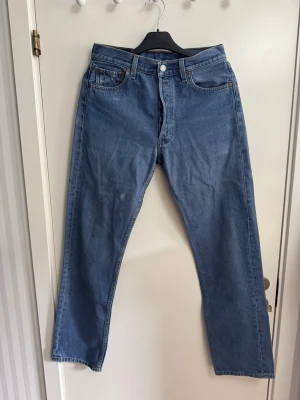 Blå raka jeans från Levi's - Säljer ett par klassiska blå jeans från Levi's med rak passform och fem fickor. Jeansen har en snygg ljusblå tvätt och är tillverkade i slitstark bomull. Perfekta för dig som gillar en tidlös och avslappnad stil. Vanliga 501. Levis lappen ser lite sliten ut men byxorna i övrigt är toppskick!