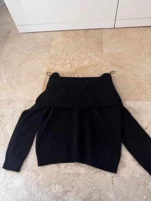 Svart off-shoulder stickad tröja - Snygg svart stickad tröja med off-shoulder design och bred ribbad krage. Tröjan har långa ärmar och ribbade muddar vid ärmslut och nederkant. Perfekt för en trendig och avslappnad look.