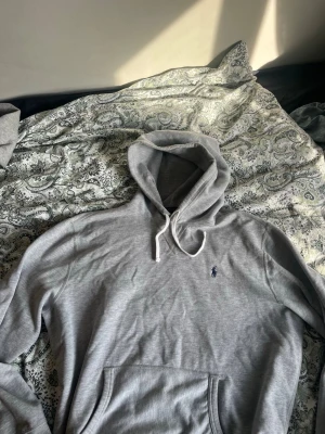 Grå hoodie från Polo Ralph Lauren - Klassisk grå hoodie från Polo Ralph Lauren med broderad logga på bröstet. Hoodien har huva med vita dragsnören och en stor magficka framtill. Tillverkad i mjukt bomullsmaterial, perfekt för en avslappnad och stilren look.
