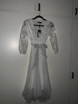 Vit långklänning med paljetter och rosett - Elegant vit långklänning från Goddiva/Bambaleno  med v-ringning, långa ärmar dekorerade med paljetter och en stor rosett i midjan. Kjolen är plisserad och har ett lätt, flytande tyg som ger en romantisk känsla. Perfekt för speciella tillfällen. Det står att storleken är (38) Men den passade mig bra , ALDRIG använt ( jag har storlek S/xs)