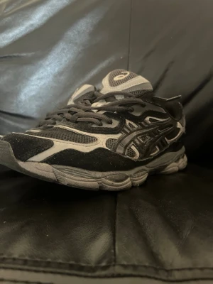 Svarta Asics sneakers med mesh och mocka - Säljer ett par svarta och grå Asics sneakers med cool design. Skorna har meshpaneler för bra ventilation och detaljer i mocka och syntet. Klassisk Asics-logga på sidan och tung design som passar streetwear. Perfekta för dig som gillar sportig och trendig stil. Priset kan diskuteras 