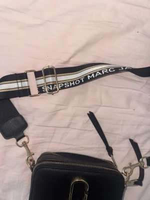 Svart axelväska Marc Jacobs Snapshot - Cool svart axelväska från Marc Jacobs Snapshot med bred justerbar axelrem i svart, vitt och beige med logotyptext. Väskan har guldfärgade detaljer och dragkedja samt en stilren, boxig form. Perfekt för att bära dina essentials med stil.
