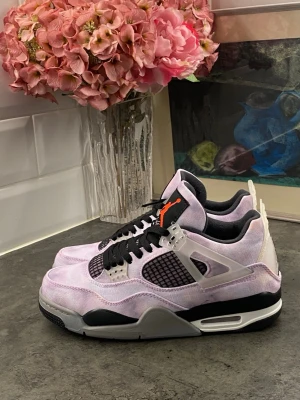 Air Jordan 4 Retro lila och svart - Snygga Air Jordan 4 Retro sneakers i lila canvas med svarta och vita detaljer. Skorna har svarta meshpaneler, svarta skosnören och en orange Jumpman-logga på plösen. Sulan är vit och svart med synlig Air-enhet. Perfekta för dig som vill sticka ut.