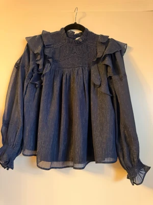 Mörkblå volangblus med glitter - En mörkblå blus med subtila glitterdetaljer och volanger på axlarna. Blusen har hög krage, långa ärmar med rynkade muddar och ett luftigt, transparent material som ger en elegant look. Perfekt för dig som gillar att sticka ut med stil.