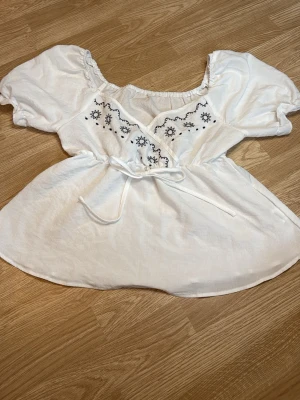 Vit blus med broderi och puffärm - Söt vit blus med blåa broderade på bröstet. Blusen har puffiga korta ärmar, en rundad halsringning med resår och knytband i midjan. I storlek S 