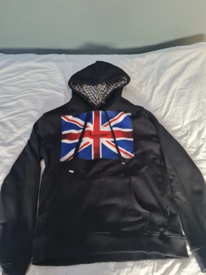 Acuascotum hoodie  - En snygg svart hoodie från aquascutum med en patch med en Storbritannien flagga, perfekt för den som vill ha en skön casual stil. 