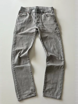 Levi’s 501 29W 32L - Snygga grå jeans i klassisk rak modell med fem fickor och normal midja. Jeansen är tillverkade i ett slitstarkt denimtyg och har en stilren look som passar till det mesta. Perfekta för dig som gillar en clean och enkel stil. En minimal fläck som syns på andra bilden.