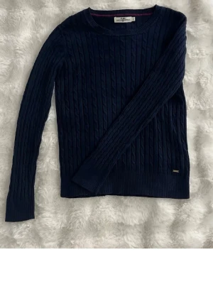 Mörkblå kabelstickad tröja - Snygg mörkblå kabelstickad tröja från Tommy Hilfiger med rund halsringning och långa ärmar. Tröjan har diskret logodetalj vid nederkanten och ribbstickade muddar. Perfekt för lager på lager och en klassisk stil.