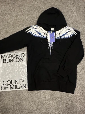Svart hoodie Marcelo Burlon med vingar - Svart hoodie från Marcelo Burlon County of Milan med ikoniskt vingtryck i vitt och blått över axlar och bröst. Klassisk huva med dragsko och känguruficka framtill. Perfekt statement-plagg för dig som gillar streetwear och unika detaljer.