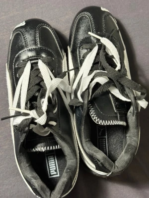 Svarta och vita Puma retro sneakers - Snygga svarta och vita sneakers från Puma med klassisk logga på sidan och tungan. Skorna har kontrasterande vita och svarta skosnören, samt detaljer i syntetmaterial och textil. Perfekta för en sportig och trendig look.