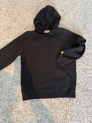 Carhartt hoodie   - Svart hoodie från Carhartt med klassisk huva och stor magficka. Storlek M.  Tröjan har ribbade muddar och en liten gul Carhartt-logga på ärmen. Perfekt för en avslappnad och stilren look. Tillverkad i mjukt bomullsmaterial. 