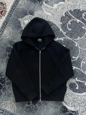 Svart hoodie från Jack & Jones - En stilren svart hoodie från Jack & Jones CORE med dragkedja framtill och huva. Hoodien har ribbade muddar vid ärmslut och nederkant samt en klassisk känguruficka. Perfekt för en avslappnad och trendig look.