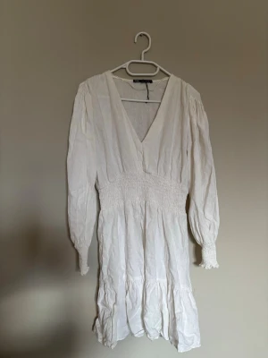Vit omlottklänning från Gina Tricot - Söt vit kortklänning från Gina Tricot med v-ringning, långa ärmar och smock i midjan. Klänningen har volangdetaljer nertill och är tillverkad i ett lätt bomullstyg. Perfekt för sommarens alla tillfällen.