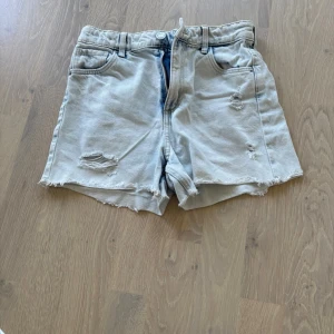 Ljusblå denimshorts med slitningar - Säljer ett par ljusblå denimshorts med råa kanter och slitningar på framsidan. Shortsen har klassisk femficksdesign, dragkedja och knapp framtill samt bälteshällor. Perfekta för sommarens alla äventyr!