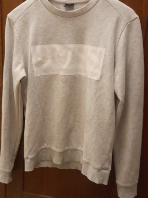 Grå sweatshirt från HUGO BOSS - Snygg grå sweatshirt från BOSS med vit logga framtill. Tröjan har rund halsringning, långa ärmar och ribbade muddar vid ärmslut och nederkant. Perfekt för en avslappnad och stilren look.