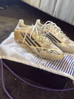 Adidas F50 fotbollsskor ELITE - Säljer ett par Adidas F50 fotbollsskor i vitt med guldiga och svarta detaljer. Skorna har snörning, låg profil och en lätt ovandel i syntetmaterial för maxad speed på planen. Ikonisk F50-logga på sidan och tå. Perfekta för dig som vill sticka ut på fotbollsplanen. Elite, därav priset