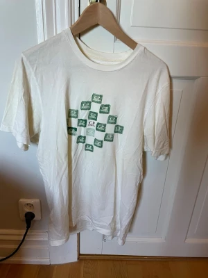 Vit t-shirt från C.P. Company - Vit t-shirt från C.P. Company med grönt tryck av loggor i rutmönster på bröstet. Klassisk rund hals och korta ärmar. Tillverkad i mjuk bomull som känns skön mot huden. Perfekt för en avslappnad och stilren look. Köpt på NK herrtrend Göteborg 