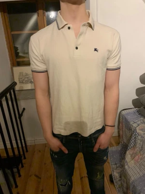 Vit pikétröja från Burberry - Klassisk vit pikétröja från Burberry med mörka detaljer på krage och ärmslut. Tröjan har korta ärmar, knappar framtill och en liten broderad logga på bröstet. Perfekt för en clean och stilren look. Den är i bra skick men saknar en knapp.