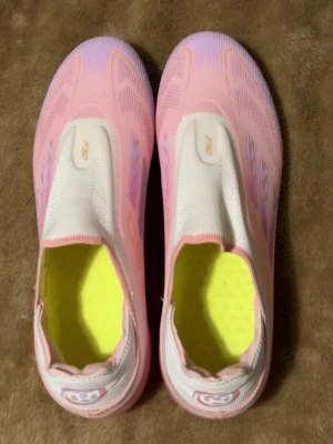 Rosa fotbollsskor från FSO - Snygga fotbollsskor från FSO i rosa och vitt med gula detaljer på sulan. Skorna har en strömlinjeformad design, elastisk ovandel och fasta dobbar för bra grepp på planen. Insidan är neonfärgad och materialet är syntetiskt för lätt vikt och komfort.