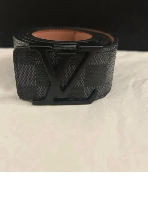 Svart rutigt LV-bälte  - Snyggt svart bälte från Louis Vuitton med klassiskt rutigt mönster och stor LV-logga i metall som spänne. Bältet är gjort i skinn och har en stilren design som passar till många outfits.