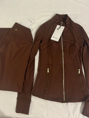 Brunt träningsset från Lululemon - Snyggt brunt träningsset från Lululemon med en figurnära jacka med dragkedja och två fickor samt matchande tights. Materialet är stretchigt och följsamt, perfekt för träning, yoga eller för vardagen. Guld detaljer. 