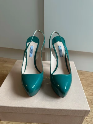 Petrol lackpumps från Prada - Snygga petrol pumps från Prada i glansigt lackläder. Skorna har öppen häl med smal rem och spetsig tå, vilket ger en elegant och trendig look. Perfekta för dig som vill sticka ut med färg och exklusiv design.