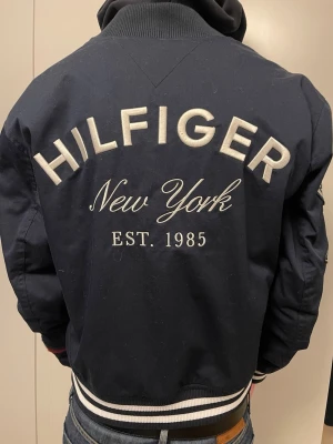 Marinblå Hilfiger bomberjacka - Snygg marinblå bomberjacka från Tommy Hilfiger med stora broderade loggor och texten 'Hilfiger New York EST. 1985' på ryggen. Jackan har vita och röda detaljer, ribbade muddar och coola patches på ärmen. Helt ny med prislapp!