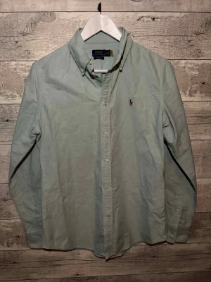 Ljusblå skjorta från Polo Ralph Lauren - Klassisk ljusblå skjorta från Polo Ralph Lauren med button-down krage och broderad logga på bröstet. Skjortan har långa ärmar, normal passform och knäppning framtill. Perfekt till jeans eller chinos för en clean look.