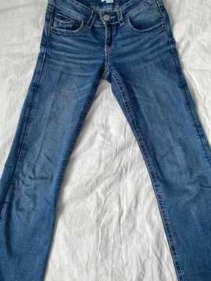 Blå bootcut jeans med låg midja - Snygga blå bootcut jeans med låg midja och klassiska fem fickor. Jeansen har en lätt tvättad look och kontrastsömmar. Perfekta för en avslappnad och trendig stil. Materialet är denim i bomull med lite stretch för extra komfort.