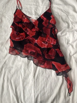 Röd blommig linnetopp med volanger - Säljer en snygg linnetopp från Pull&Bear med röda blommor på svart botten. Toppen har tunna axelband, volangdetaljer och asymmetrisk nederkant. Perfekt för dig som vill sticka ut lite extra med ett unikt mönster och luftig känsla.