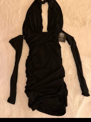 Svart kort klänning med halterneck - Snygg svart kort klänning från HEY med halterneck och åtsittande passform. Klänningen har rynkade detaljer längs sidorna och är ärmlös, perfekt för en trendig look.