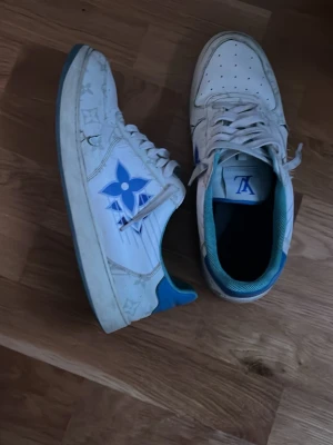 Louis Vuitton vita och blå sneakers - Säljer ett par vita Louis Vuitton sneakers med blå detaljer och ikoniska LV-monogram på sidorna. Skorna har lågt skaft, perforerad tå och blå insida. Tillverkade i läder med klassisk snörning och gummisula. Perfekta för dig som vill sticka ut med exklusiv stil.