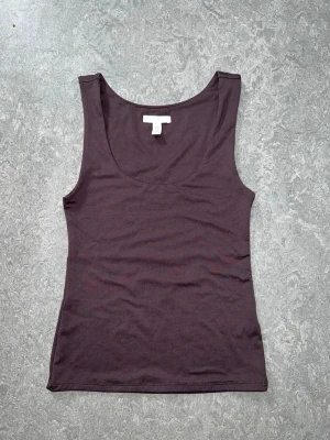 Mörkbrun ärmlös linnetopp - En enkel och stilren mörkbrun linnetopp med rund halsringning. Toppen är ärmlös och har en figurnära passform, perfekt att matcha med jeans eller kjol. Tillverkad i mjuk bomull för en bekväm känsla. Den har även dubbeltyg.