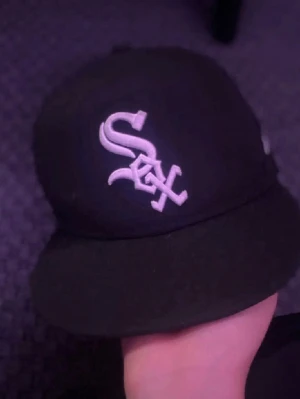 Svart White Sox keps New Era - Säljer en svart keps från New Era med White Sox-logga broderad i vitt framtill och MLB-logga bak. Klassisk rak skärm och stilren design, perfekt för dig som gillar sportiga accessoarer och streetwear.