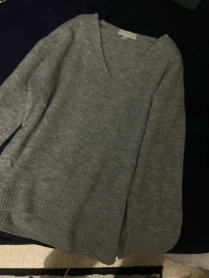 Grå stickad V-ringad tröja H&M - Mysig grå stickad tröja från H&M med V-ringning och långa ärmar. Tröjan har en avslappnad passform och ribbade muddar vid ärmslut och nederkant. Perfekt för lager på lager och enkel att matcha med jeans eller kjol.