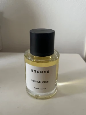 ESSNCE Sugar Kiss Eau de Parfum - ESSNCE Sugar Kiss Eau de Parfum i en stilren, rund glasflaska med svart lock. Parfymen har en ljusgul vätska och minimalistisk etikett med svart text. Perfekt för dig som gillar söta och fräscha dofter.