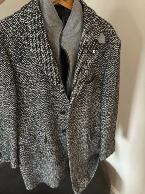 Svartvit mönstrad rock med grå väst - Stilren svartvit rock med fiskbensmönster och klassisk krage. Rocken har tre knappar framtill, fickor och en grå innerdel som liknar en väst med dragkedja. Perfekt för dig som gillar en lite varmare och längre rock. Materialet känns som ullblandning. Ny pris 5000