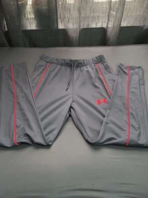 Grå mjukisbyxor från Under Armour - Snygga grå mjukisbyxor från Under Armour med röda detaljer och logga på benet. Byxorna har resår i midjan med dragsko och röda sömmar längs benen för en sportig look. Perfekta för chill eller träning.