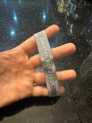 Grått armband från Dior - Snyggt grått tygarmband från Dior med texten 'Christian Dior J'Adior' broderad i vitt. Armbandet har justerbar knytning med tofsar i ändarna och är perfekt för att ge din outfit en exklusiv touch.