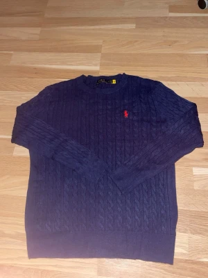 Mörkblå kabelstickad tröja Polo Ralph Lauren - Mörkblå kabelstickad tröja från Polo Ralph Lauren med klassisk rund halsringning och röd broderad logga på bröstet. Tröjan har ribbade muddar vid ärmslut och nederkant. Perfekt för dig som gillar stilren och tidlös design.