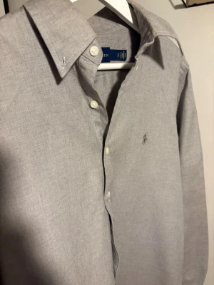 Grå skjorta från Ralph Lauren - Klassisk grå (med gröna toner) skjorta från Ralph Lauren med diskret logobrodyr på bröstet. Skjortan har lång ärm, knappar framtill och en stilren, tidlös design. Tillverkad i mjuk bomull som känns skön mot huden. Perfekt för dig som gillar enkel och clean stil.