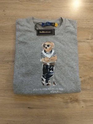Grå sweatshirt Polo Bear Ralph Lauren - Cool grå sweatshirt från Ralph Lauren med Polo Bear-tryck på framsidan. Tröjan har rund hals, ribbade muddar och är tillverkad i mjukt material. Perfekt för dig som gillar klassisk streetstyle och ikoniska prints. Det står att den är Large men den är i storleken Small