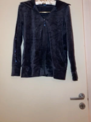 Svart velourhoodie med dragkedja - Mjuk och skön svart hoodie i velourmaterial med dragkedja framtill och huva. Långa ärmar och breda muddar vid ärmslut och nederkant. Perfekt för en avslappnad och trendig look.