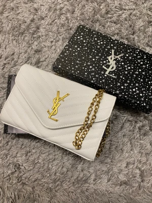 Vit YSL kuvertväska med guldkedja - Snygg vit kuvertväska från YSL i läder med klassiskt YSL-emblem i guld framtill. Väskan har quiltat mönster och en elegant guldfärgad kedja som axelrem. Perfekt accessoar för att lyfta din outfit.