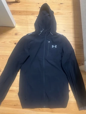 Svart vindjacka från Under Armour - Svart vindjacka med huva från Under Armour. Jackan har dragkedja framtill, reflekterande detaljer på axlarna och logga på bröstet. Perfekt för blåsiga dagar och sportiga outfits.