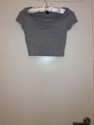 Grå croppad topp med kort ärm - Snygg grå croppad topp med korta ärmar och lätt rynkad detalj vid urringningen. Tillverkad i mjukt bomullsmaterial som känns skönt mot huden. Perfekt till jeans eller kjol för en avslappnad look.
