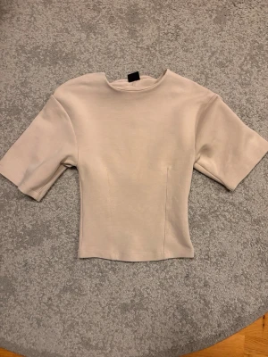 Beige topp från Gina Tricot - Snygg beige topp från Gina Tricot med korta ärmar och rund halsringning. Modellen är figurnära och har en stilren, enkel design som passar till många olika looks. Tillverkad i mjukt material för en bekväm känsla.